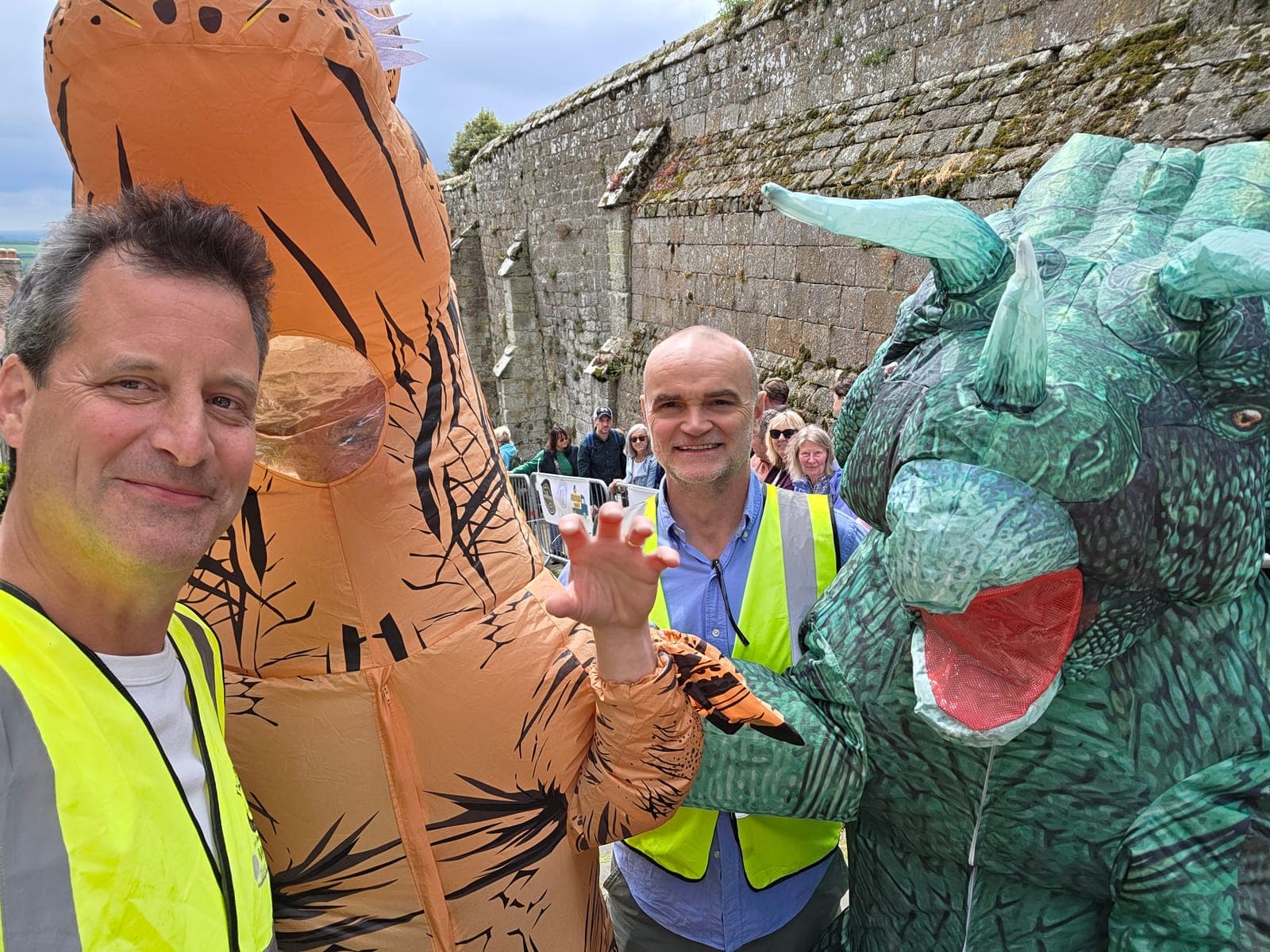 Jim Chetwode, left, in a high-vis vest beside a fellow marshal and two competitors in inflatable dinosaur costumes, in front of the stone wall on Gold Hill in Shaftesbury.